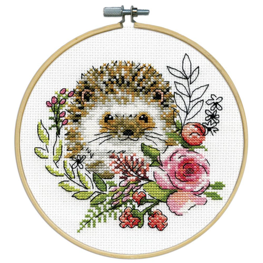 A beginner-friendly Design Works counted cross-stitch kit featuring a cute hedgehog design. The kit includes 11-count white Aida fabric, a pre-sorted 100% cotton thread palette, a tapestry needle, and an 8-inch circular hoop. The packaging highlights the whimsical, kid-friendly illustration and the all-in-one convenience of the set.