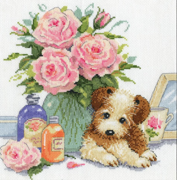 An adorable Design Works counted cross-stitch kit titled 'Puppy with Rose.' The design features a realistic, fluffy puppy with a gentle expression, framed by soft pink roses and green leaves in the background. The kit is shown with 14-count white 100% cotton Aida cloth, a thread sorter with pre-sorted cotton floss, a needle, and instructions