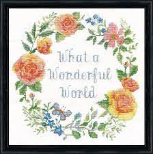 A Design Works counted cross-stitch kit titled 'Wonderful World.' The design features a circular garland of yellow and orange roses with green leaves and butterflies, surrounding the hand-scripted text 'What a Wonderful World' in light blue. The kit includes 14-count white Aida fabric, a pre-sorted thread palette of 100% cotton floss, a needle, and instructions
