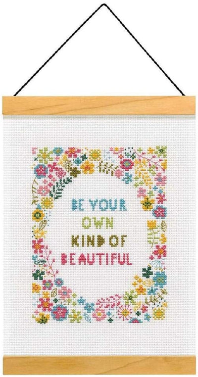 An elegant Dimensions counted cross-stitch kit featuring a large bouquet of detailed flowers in pinks, purples, and greens surrounding the inspirational quote 'Be Your Own Kind of Beautiful.' The kit includes 14-count white Aida, embroidery thread, a needle, and two wooden strips for a decorative hanging finish