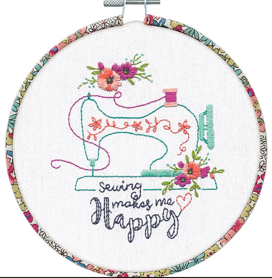 A charming Dimensions embroidery kit titled 'Sew Happy.' The design features the phrase 'Sewing Makes Me Happy' in a whimsical font, surrounded by colorful thread spools, needles, and floral accents. The kit includes fabric with the design pre-printed on it, a 6-inch bamboo hoop, pre-sorted embroidery thread, and a needle