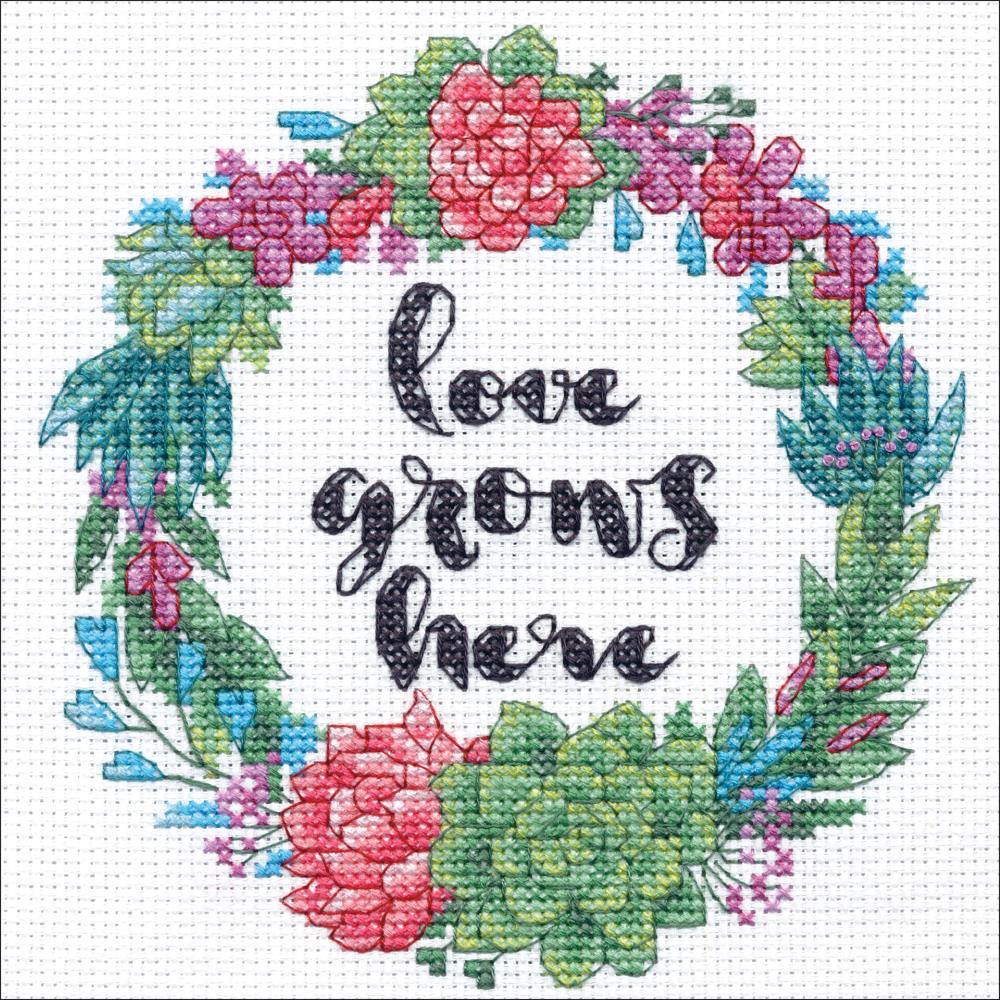 A charming Dimensions counted cross-stitch kit featuring a wreath of various succulents in shades of sage, mint, and dusty rose. In the center, the phrase 'Love Grows Here' is stitched in elegant script. The kit includes 14-count white Aida cloth, cotton embroidery thread, a needle, and instructions for a 6-inch square finished piece.