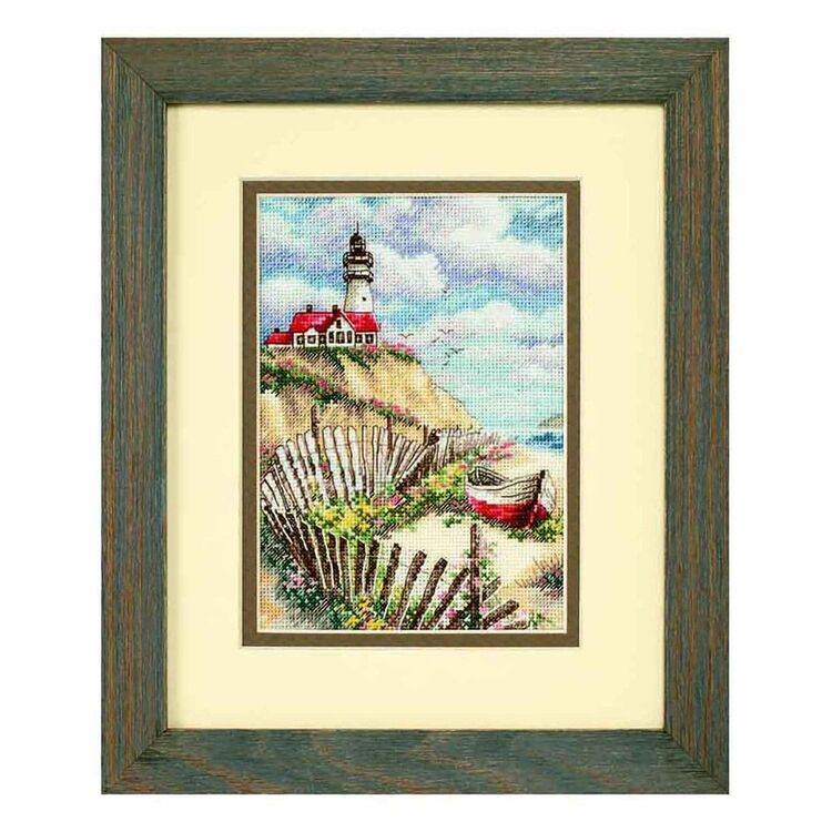 A framed cross stitch kit depicting a lighthouse on a coastal background, within a brown frame.