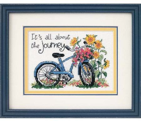 An inspirational Dimensions counted cross-stitch kit featuring a vintage bicycle with a flower basket and the quote 'It's all about the journey.' The design uses a soft, inviting color palette. The kit includes 14-count white Aida fabric, pre-sorted embroidery threads, a needle, and a clear pattern sheet for a 5x7 inch finished piece.