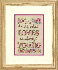 A vibrant Dimensions embroidery kit designed by Lori Siebert. The design features an abundance of colorful, stylized flowers in shades of pink, orange, turquoise, and yellow surrounding an inspirational Greek proverb. The artwork is pre-printed on a warm beige fabric background. The kit includes pre-sorted cotton thread, a needle, and easy instructions for a 5x7 inch finished piece."