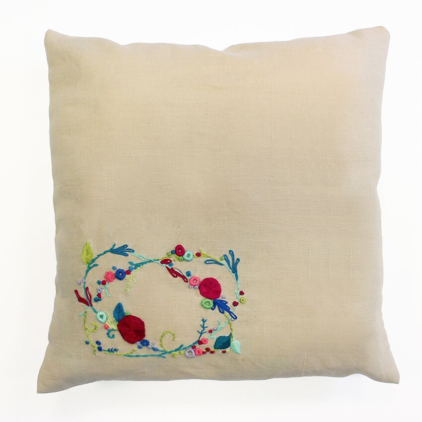 A cushion cover with a floral embroidery design in the center, featuring a rose garland motif.
DMC Sweet Meadow Rose Garland embroidery kit packaging, showing a stunning finished floral wreath cushion on a rustic chair.