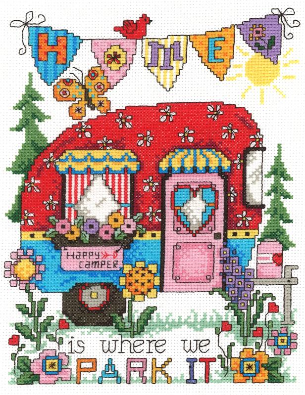 A vibrant counted cross-stitch kit titled 'Happy Camper' by designer Diane Arthurs. The design features a whimsical, retro-style travel trailer surrounded by outdoor motifs in a bright, illustrative style. The kit includes 14-count white Aida fabric, premium DMC embroidery threads, and a needle. The finished design measures 8.7 inches wide by 6.7 inches high