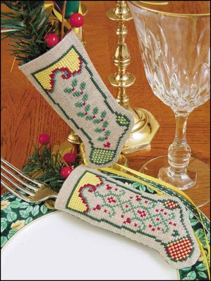 Two cross-stitched linen stockings with a festive design including green and red patterns, displayed on a table setting with a glass and Christmas decorations.