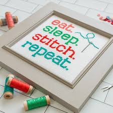 A square-format cross-stitch kit from It’s Sew Emma titled 'Eat Sleep Stitch Repeat.' The design features a modern, typographic layout of the popular maker's motto, surrounded by whimsical stitching motifs like scissors, thread, and needles in a bright, contemporary palette. The kit includes 14-count white Aida cloth, full skeins of DMC embroidery threads, and a needle. The design measures 70 stitches wide by 70 stitches high.