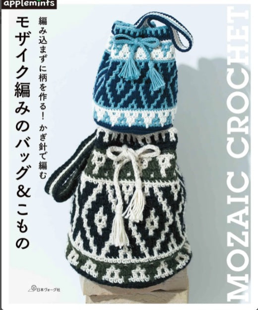 The cover of the book 'Mozaic Crochet' displays two crocheted bags, one in black and white with a blue trim, and the other in green with a white pattern. Japanese text is visible on the cover, suggesting it is written in Japanese. Front cover of a Japanese Mosaic Crochet book featuring a bold, geometric patterned blanket in contrasting colors