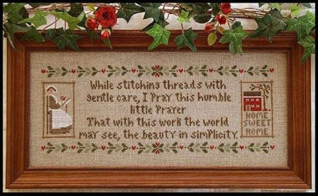 The 'A Stitcher's Prayer' cross-stitch sampler featuring traditional primitive motifs and a heartwarming poem about the beauty of stitching.
The finished 'A Stitcher's Prayer' framed in a weathered wood frame, hung in a cozy, sunlit sewing room