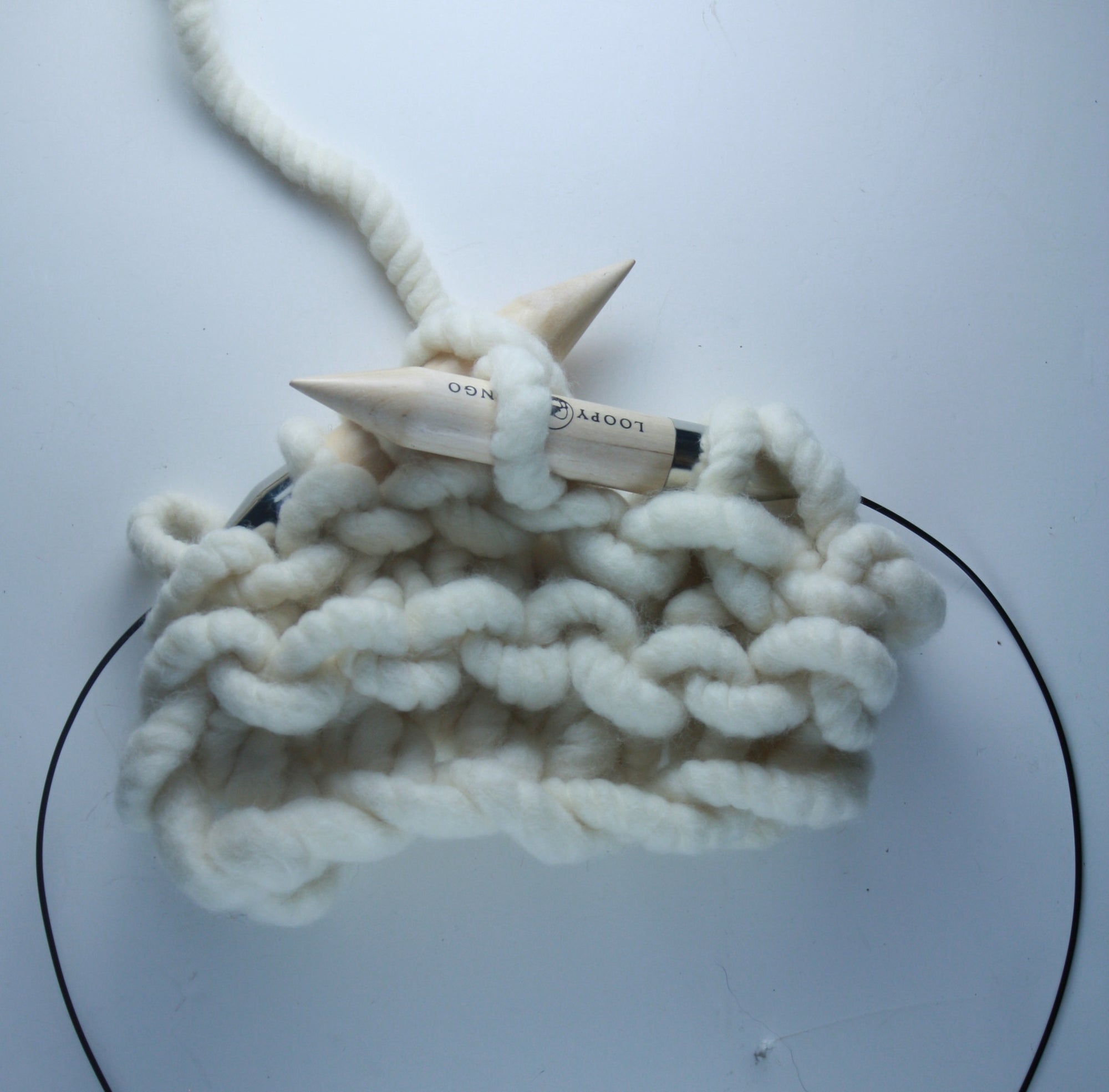 A pair of giant circular knitting needles made from birch wood, with a flexible plastic connecting wire, resting on a knitted fabric.

25mm giant circular needles coiled next to a ball of chunky yarn, demonstrating their compact, travel-friendly size
