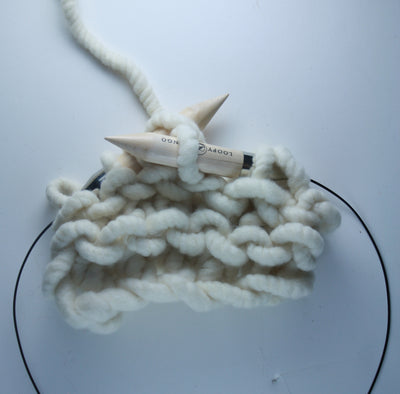 A pair of giant circular knitting needles made from birch wood, with a flexible plastic connecting wire, resting on a knitted fabric.

25mm giant circular needles coiled next to a ball of chunky yarn, demonstrating their compact, travel-friendly size