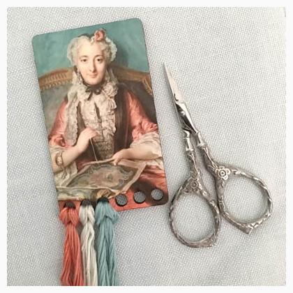 A wooden thread holder with a vintage postcard design featuring an illustration of a woman in a pink and blue dress, alongside a pair of silver-handled scissors.