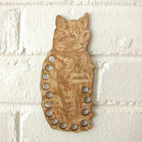 A wooden thread organizer in the shape of a cat with the word 'stitch' inscribed, featuring holes for organizing floss.
