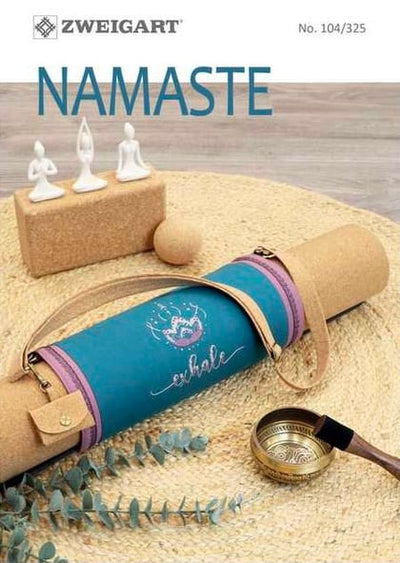 A cross stitch pattern book with a blue and brown cover, labeled 'NAMASTE', with a small decorative figure and a tealight candle on the foreground.
