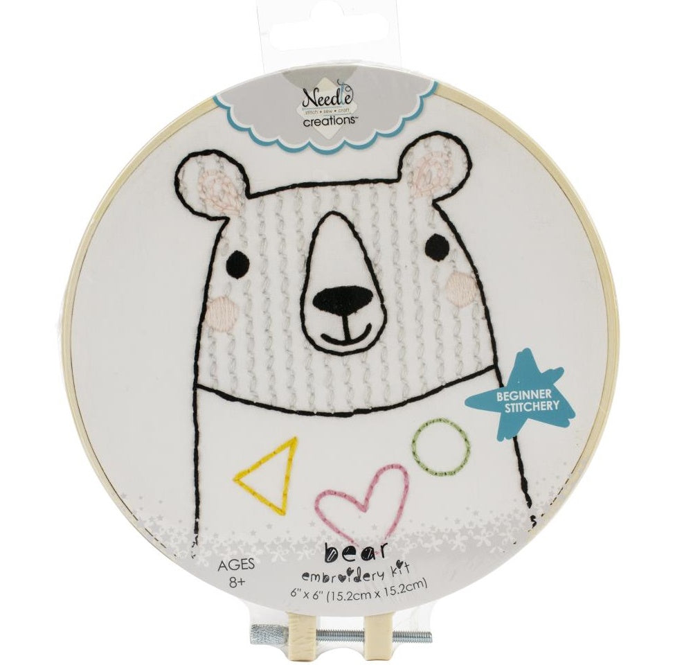 A circular beginner's embroidery kit from Needle Creations' Stitchin' Kidz line featuring a cute, whimsical bear design. The design is pre-printed on a soft felt fabric panel, making it easy for beginners to follow the lines. The kit includes a 6-inch plastic embroidery hoop, colorful embroidery floss, a metal needle, and step-by-step instructions. The finished project is displayed within the colorful hoop