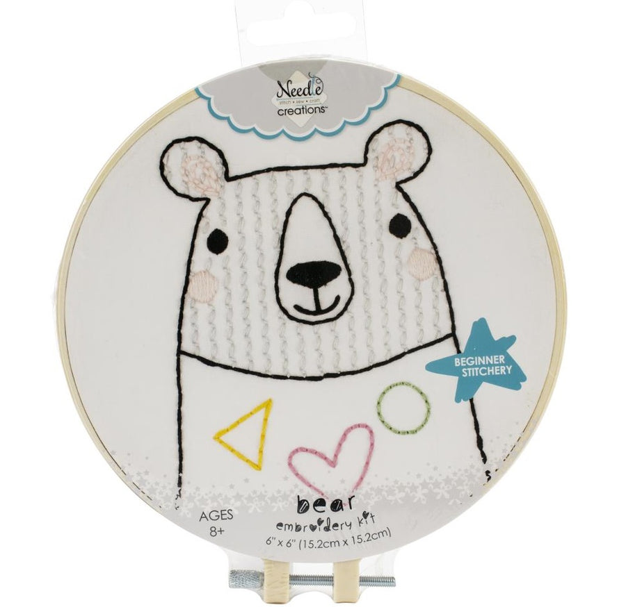 A circular beginner's embroidery kit from Needle Creations' Stitchin' Kidz line featuring a cute, whimsical bear design. The design is pre-printed on a soft felt fabric panel, making it easy for beginners to follow the lines. The kit includes a 6-inch plastic embroidery hoop, colorful embroidery floss, a metal needle, and step-by-step instructions. The finished project is displayed within the colorful hoop