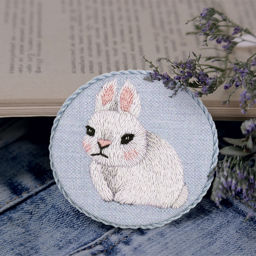 A specialized miniature embroidery kit by Panna for creating a circular Baby Rabbit brooch. The kit features fabric with a pre-printed bunny design, a curated palette of soft grey and white embroidery threads for realistic fur texture, a needle, and brooch hardware. The finished 6.5cm accessory displays a sweet, detailed rabbit face with expressive eyes