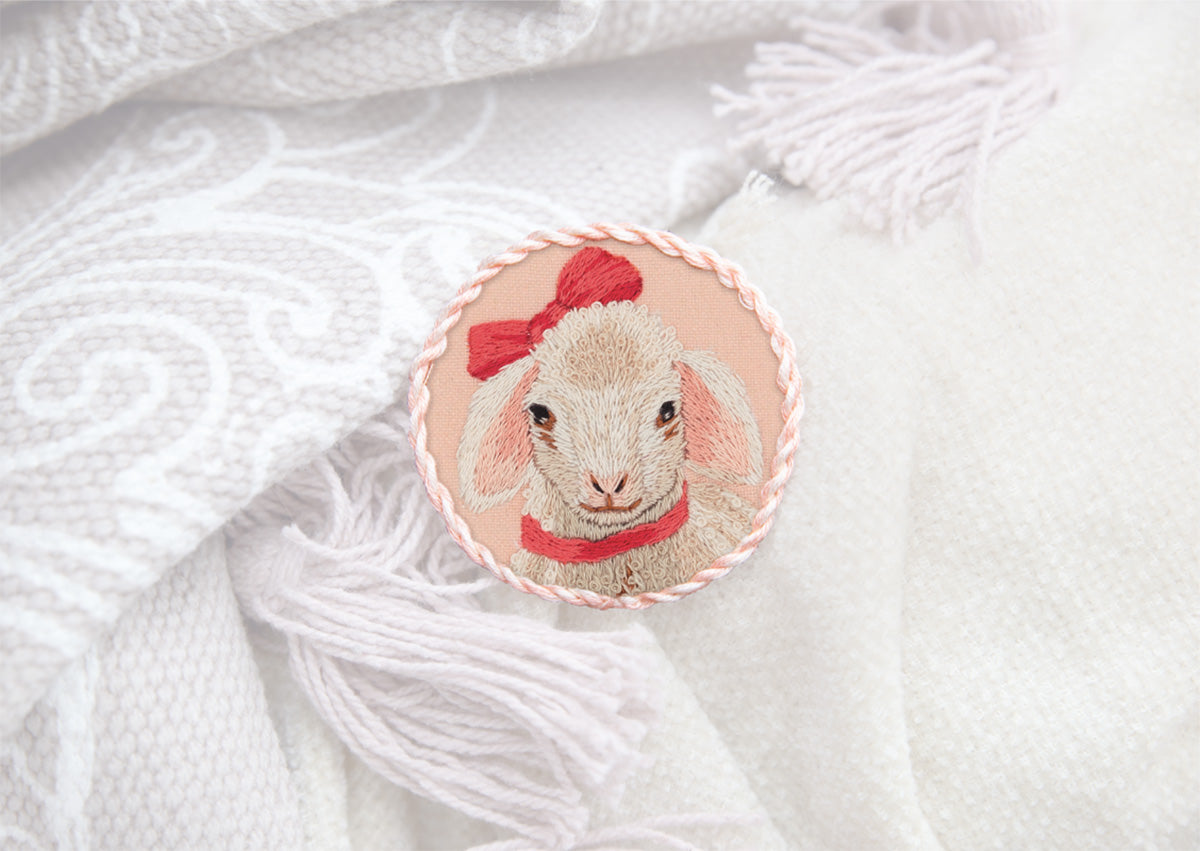 A hand-embroidered brooch with a lamb design on a white fabric background, resembling a patch that is detached from a larger garment.
