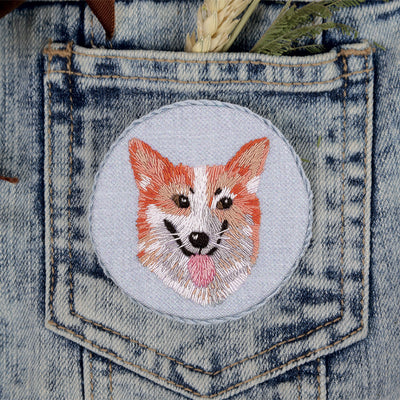 A specialized embroidery kit by Panna for creating a circular Welsh Corgi brooch. The kit features fabric with a pre-printed corgi design, a curated selection of embroidery threads to mimic realistic fur, a needle, and a brooch back component. The finished 6.5cm accessory displays a charming, detailed portrait of a corgi in warm tan and white tones