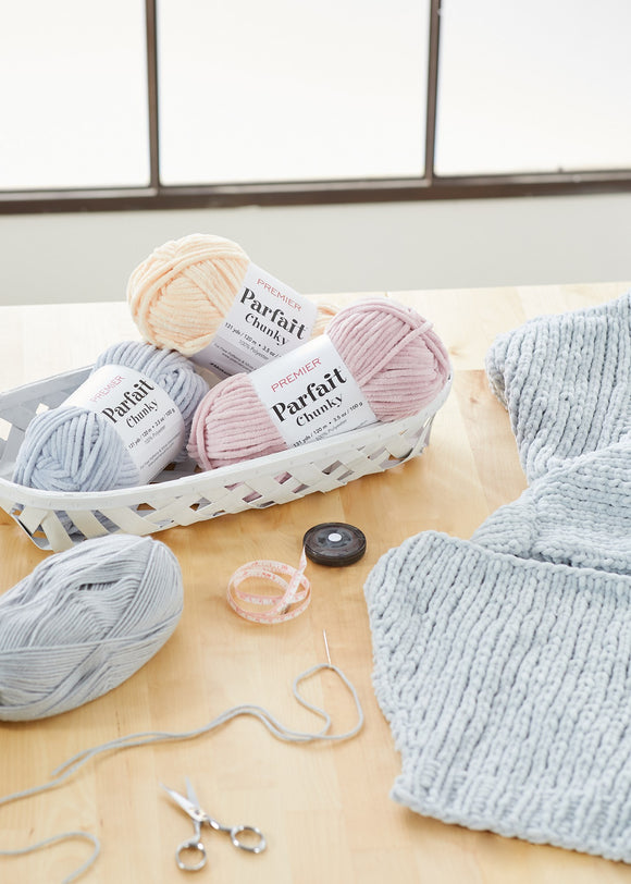 Several skeins of Premier Parfait chunky yarn in gray and pink colors are displayed alongside a partially crocheted fabric piece on a table.