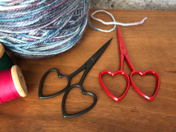 A pair of 3.75-inch 'Red Hots Love' embroidery scissors featuring elegant heart-shaped handles. The scissors are shown in two vibrant color options, highlighting their sharp, tapered blades and polished finish, designed for precise thread cutting in cross-stitch and quilting.
