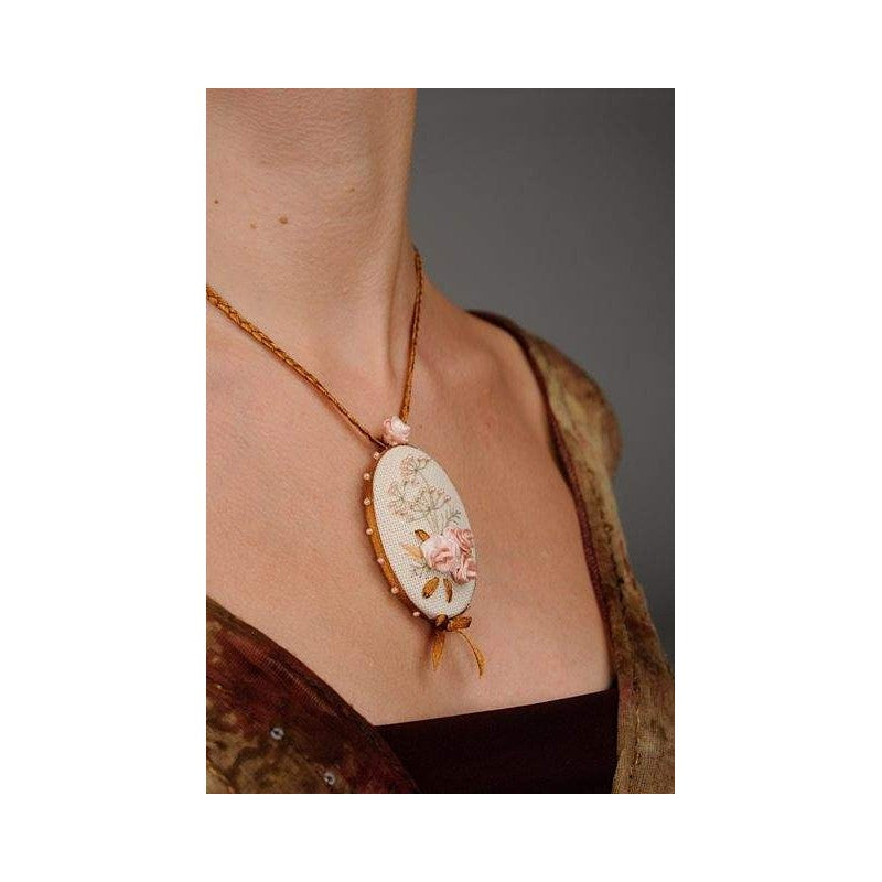 An elegant wearable embroidery kit from Riolis for creating a rose-themed pendant. The design features a delicate rose crafted with silk ribbons, accented by Anchor embroidery floss and shimmering beads. The kit includes high-quality 25-count Zweigart Lugana fabric, a decorative metal pendant frame, two specialized needles, and full instructions. The finished oval-shaped pendant measures 4.5cm by 6.5cm