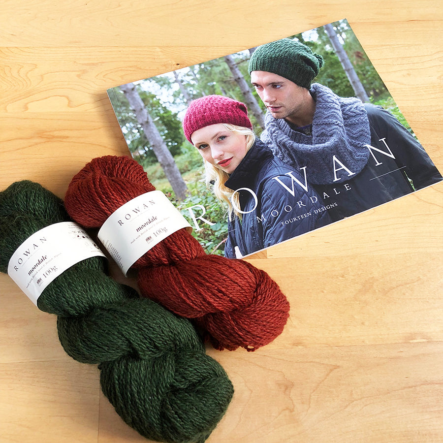 2 skeins of Rowan Moordale yarn in red and pine green colors, alongside a knitting needle and a pattern book with a couple wearing knitted hats on the cover.