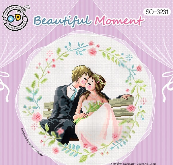 A romantic and whimsical cross-stitch kit from SODA (Korea) titled 'Beautiful Moment.' The design features an illustrative and decorative layout with floral elements and romantic symbols in a soft, elegant color palette. The kit includes 14-count white Aida cloth, full skeins of DMC embroidery threads, and a needle. The finished design measures 22 cm wide by 21.5 cm high.