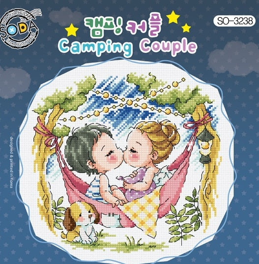 A charming, square-format cross-stitch kit from SODA (Korea) titled 'Camping Couple.' The design features a whimsical illustration of a couple in a campsite setting, surrounded by a tent, pine trees, and a campfire in a vibrant, modern color palette. The kit includes 14-count white Aida cloth, full skeins of DMC embroidery threads, and a needle. The finished design measures 19 cm wide by 18 cm high