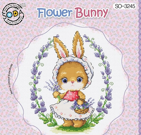 A cross stitch kit image featuring a whimsical brown bunny with a floral dress and a bonnet, sitting beside a basket of purple flowers, on a white background with a pink floral border.