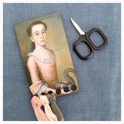 A wooden thread holder in the shape of a vintage postcard, featuring an illustration of a schoolgirl holding a squirrel, with thread compartments in the background. A pair of scissors is also present to the side.