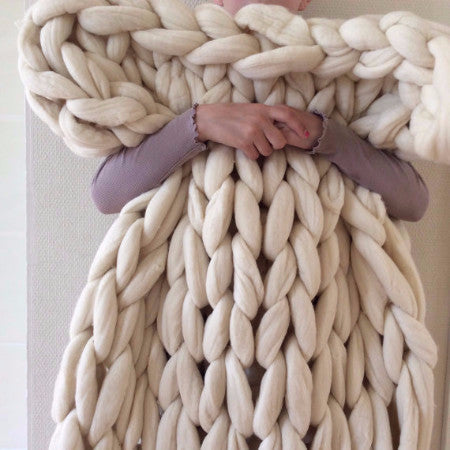 A person holding a large, chunky, off-white merino wool blanket yarn.

"A finished, giant-knit Merino blanket in a modern living room, showcasing the heavy drape and cozy, oversized stitches.
