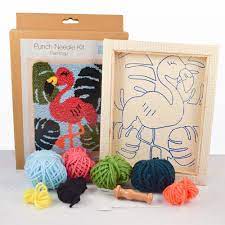 The kit includes an 8x10 inch fabric panel pre-stretched on a wooden frame with a printed flamingo silhouette. It features a palette of vibrant pink and coral yarns, a specialized punch needle tool, a needle threader, and a large color image for easy reference