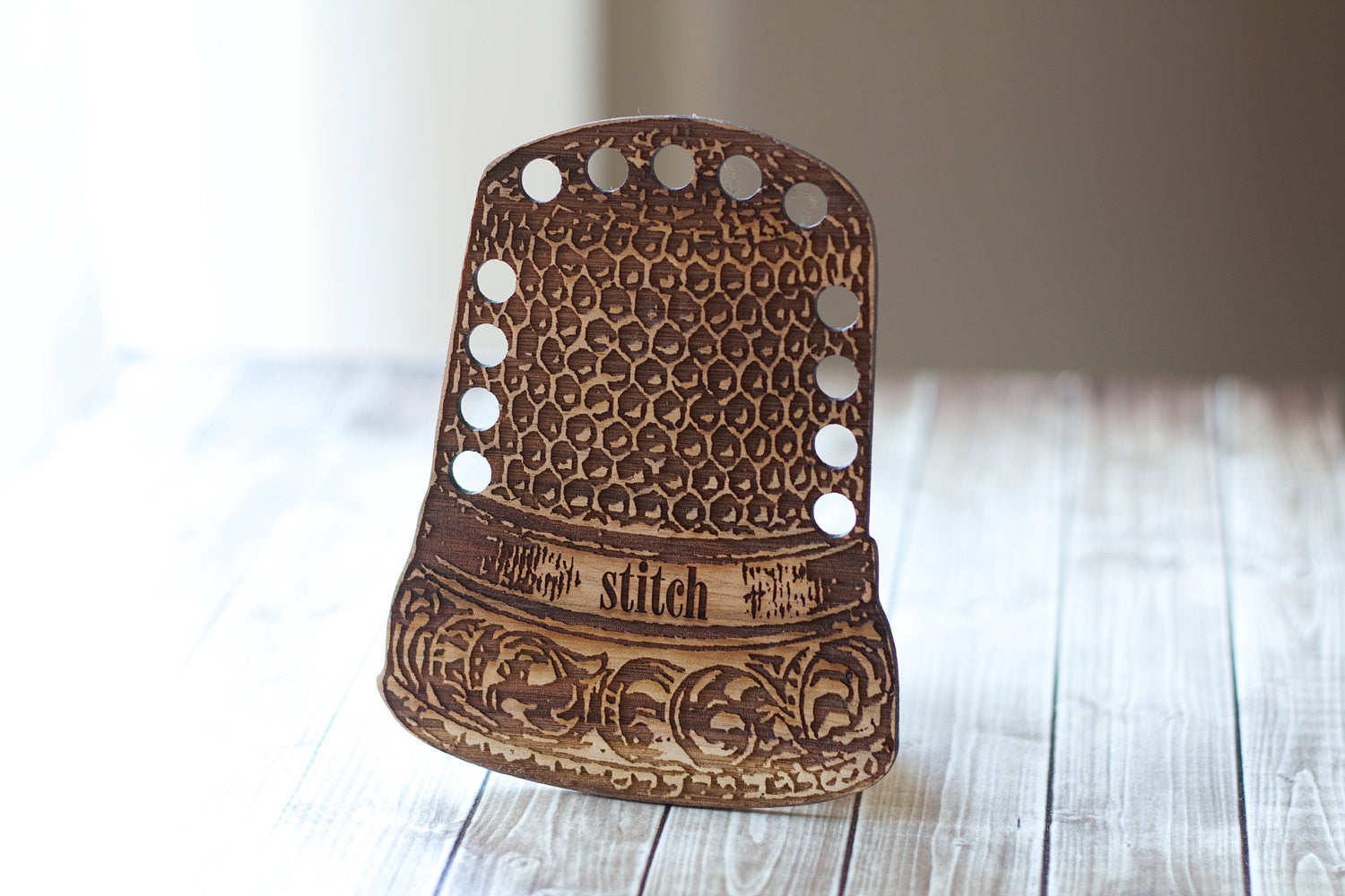 A professional-grade wooden floss organizer shaped like a classic vintage tailor's thimble. Crafted in the USA from premium local hardwoods, this high-definition tool features 13 precision-drilled holes for thread organization. The surface is adorned with a charming, laser-etched dimple pattern and vintage scrollwork that mimics a traditional metal thimble.