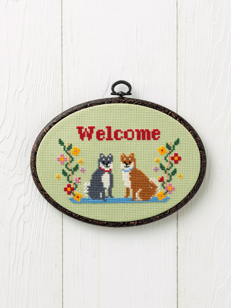 An all-in-one cross-stitch kit imported from Japan featuring a 'Dog' welcome design. The kit is shown with its 14-count cotton stitching cloth, vibrant 100% cotton threads, and a custom 14cm x 19cm frame. The image highlights the kid-friendly illustrative pattern and the high-quality materials included for an immediate start.