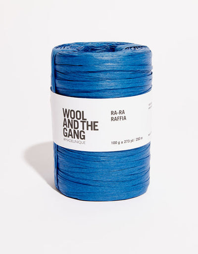A roll of riviera blue Wool And The Gang Ra-Ra Raffia yarn with a label in the center.