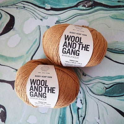 Two skeins of Buddy Hemp Yarn in beige color placed on a textured blue and green surface.
A 100g ball of Wool and the Gang Buddy Hemp yarn, showcasing its natural, matte texture and soft, earthy color palette