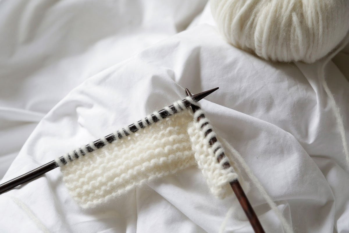 A close-up image of white yarn being knitted with wooden knitting needles, with a skein of yarn visible in the background.

"Close-up of Feeling Good yarn, highlighting the delicate Baby Alpaca fibers held within a fine cage structure for a weightless, cloud-like feel.