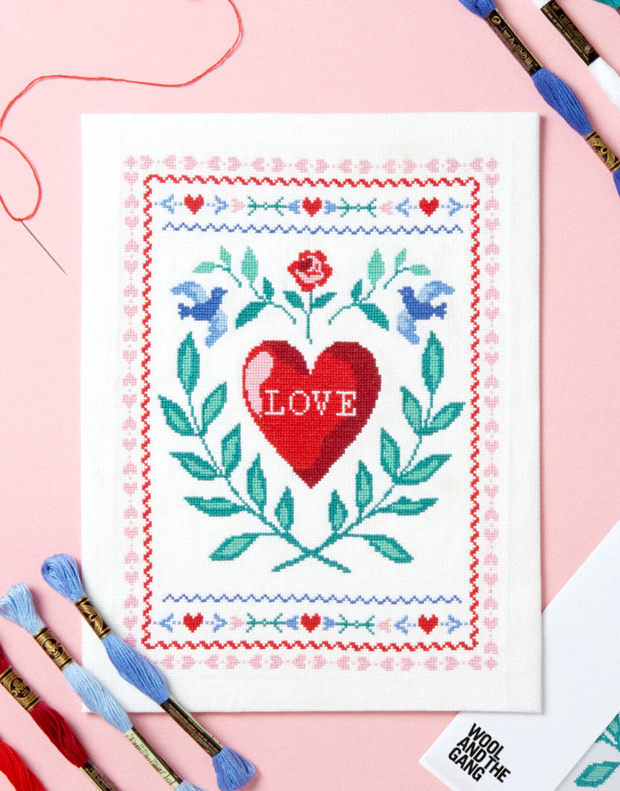 Cross stitch kit packaging with a completed design featuring a red heart with the word 'LOVE' and a floral border, surrounded by Embellishments such as embroidery threads and needles.