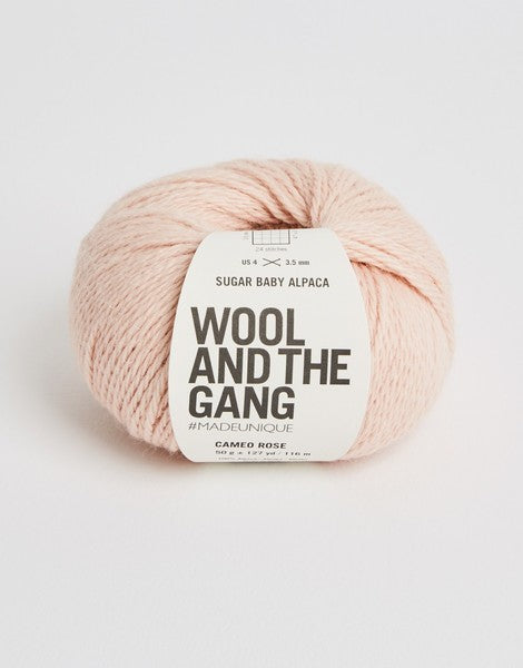 A blue skein of WOOL AND THE GANG Sugar Baby Alpaca yarn with a label in the center in cameo rose colour