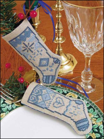 Two cross-stitched linen stockings with a blue and green pattern, displayed on a table setting.