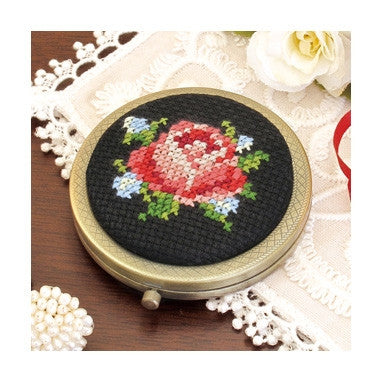 A compact mirror with a cross stitch design on the cover, featuring a floral pattern primarily in red and blue colors, framed in a metallic gold border.